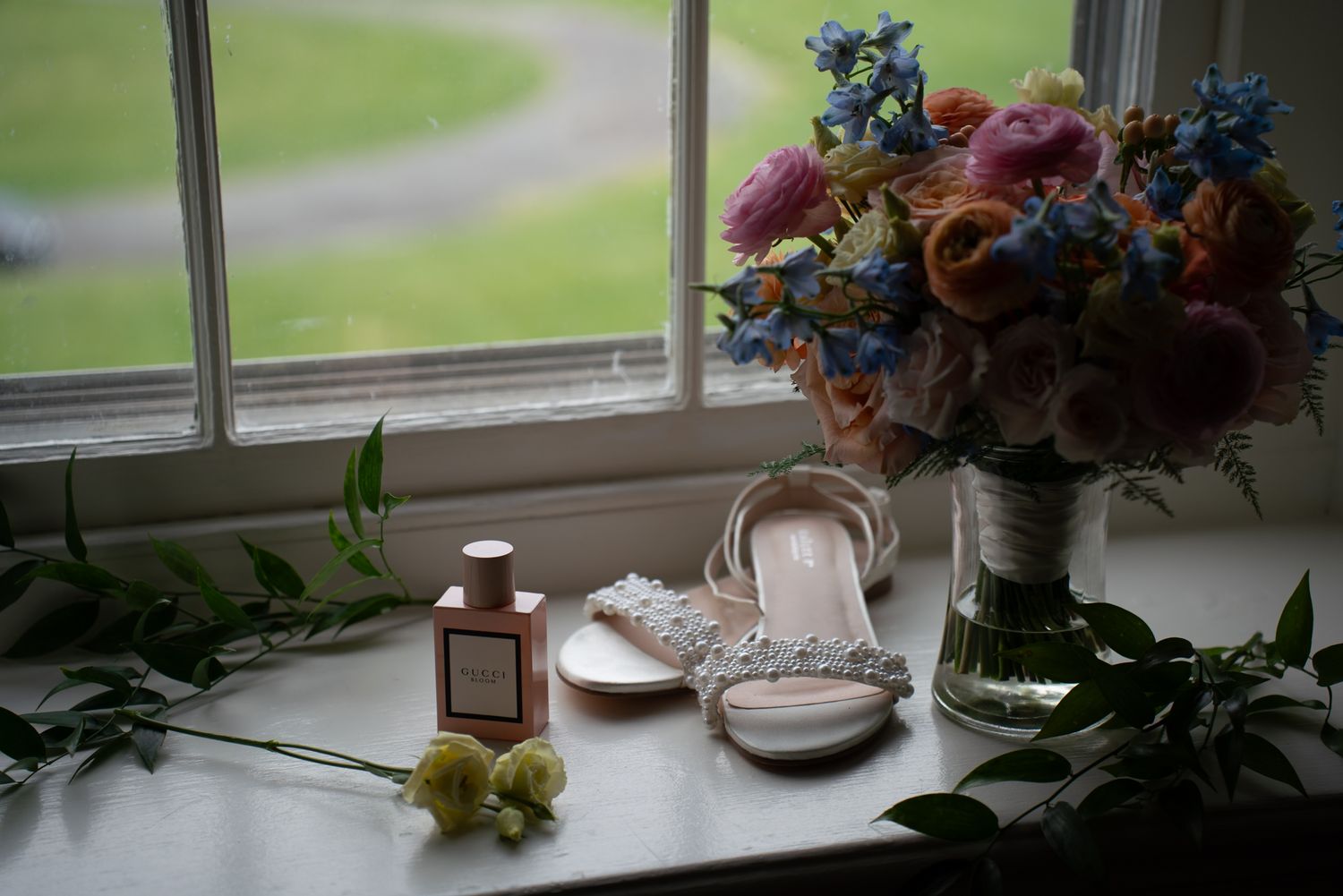 Bridal flat shoes, Gucci perfume, and colorful wedding bouquet with blue and pink flowers on a windowsill.