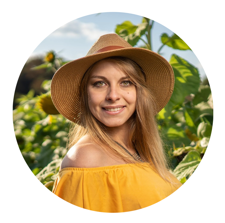 Smiling blonde woman in yellow off-shoulder top and straw hat standing in a sunflower field.