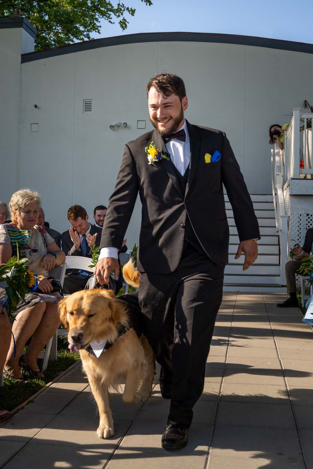 Groom in black tuxedo walks golden retriever in tuxedo down the aisle at outdoor wedding ceremony.