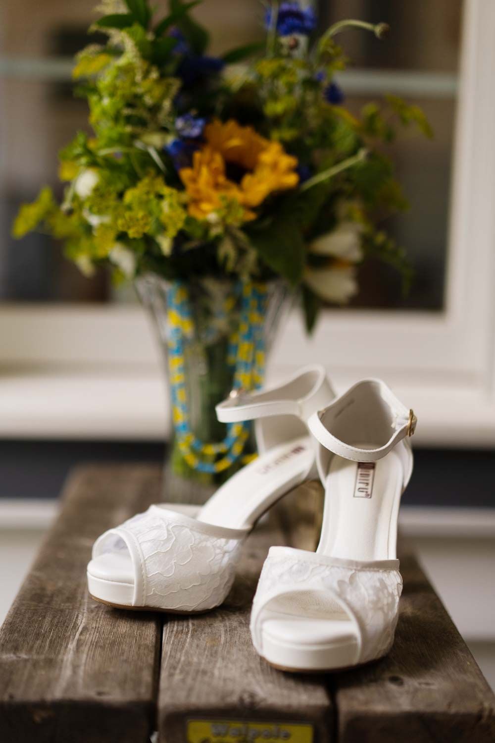 White lace bridal heels displayed on rustic wood surface with colorful sunflower bouquet behind.