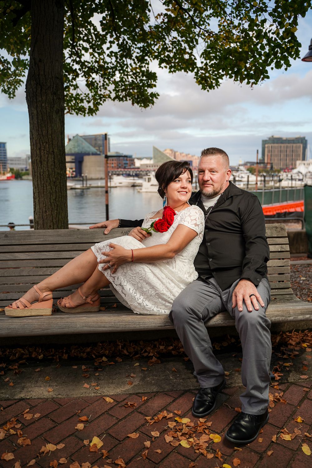 Wedding couple sitting on a park bench by the harbor, bride in white lace dress holding red roses, groom in black shirt.