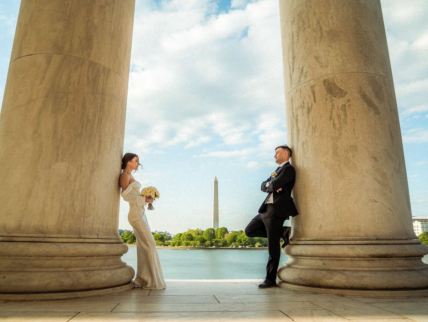 Bride and groom stand between marble columns with the Washington Monument reflecting in the Tidal Basin.