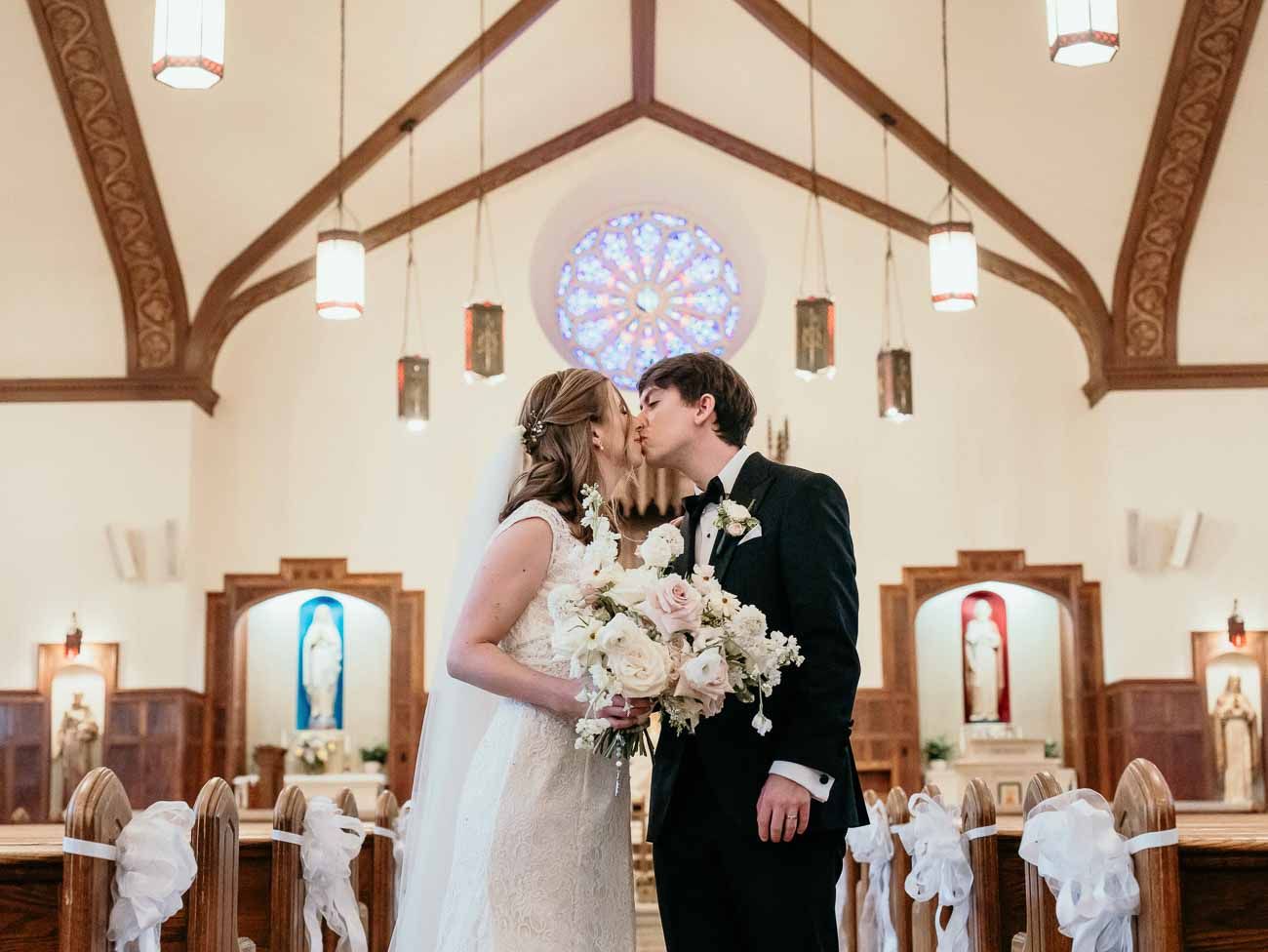 Bride and groom share a kiss at the altar of an elegant church with a rose window and white decor.