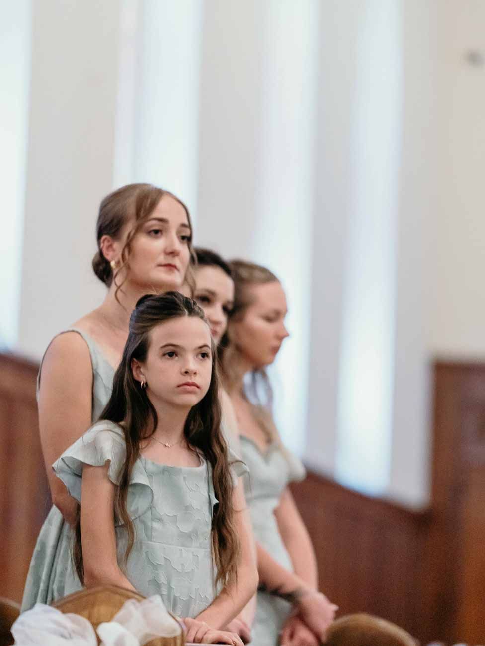 Bridesmaids in sage green dresses seated attentively in church pews during wedding ceremony.