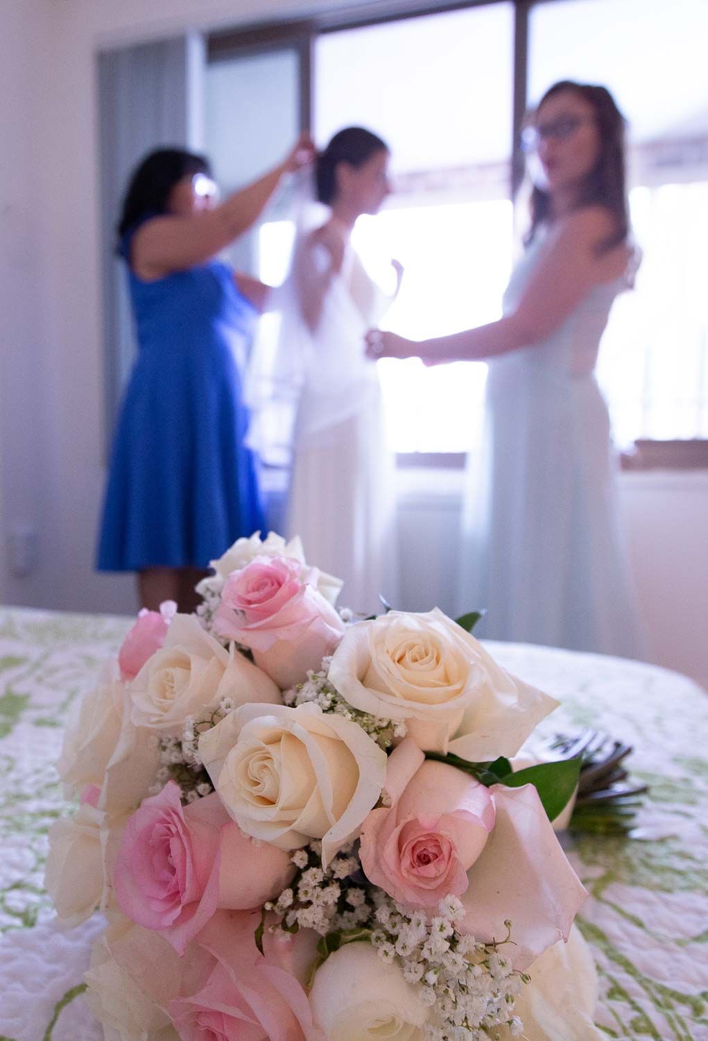 Beautiful bridal bouquet of pink and white roses in foreground with bridesmaids helping bride dress by window.