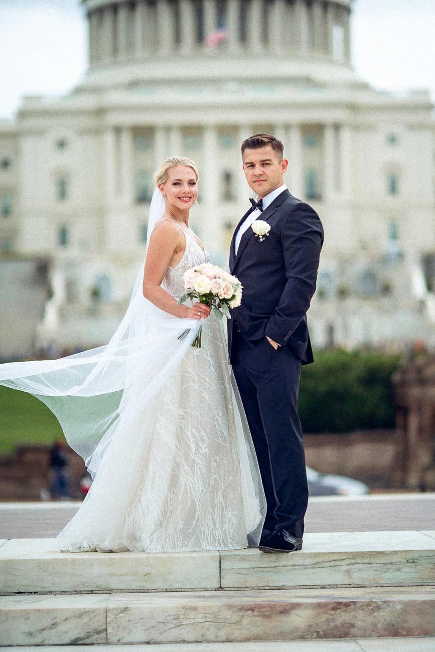 Bride and groom posing on steps in front of the US Capitol building, bride holding white bouquet with veil flowing.