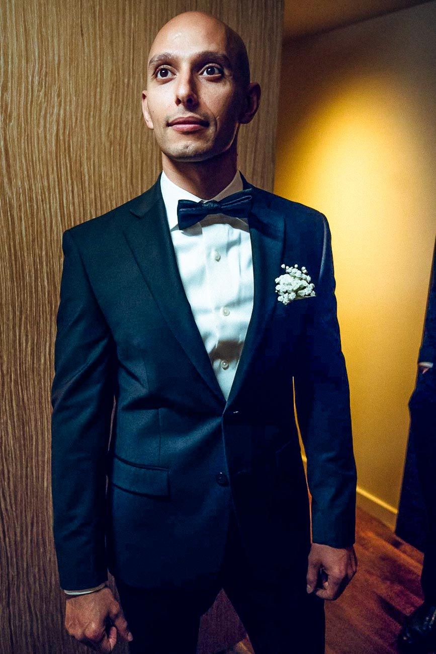 Bald groom in black tuxedo with bow tie and boutonniere posing indoors before wedding ceremony.
