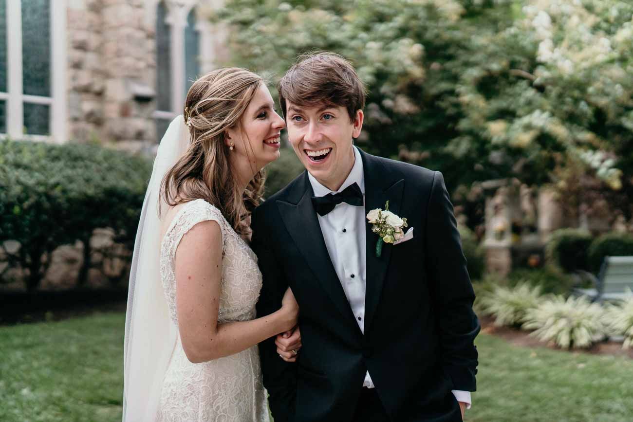 Laughing bride and groom share a joyful moment together outdoors near a stone church building.