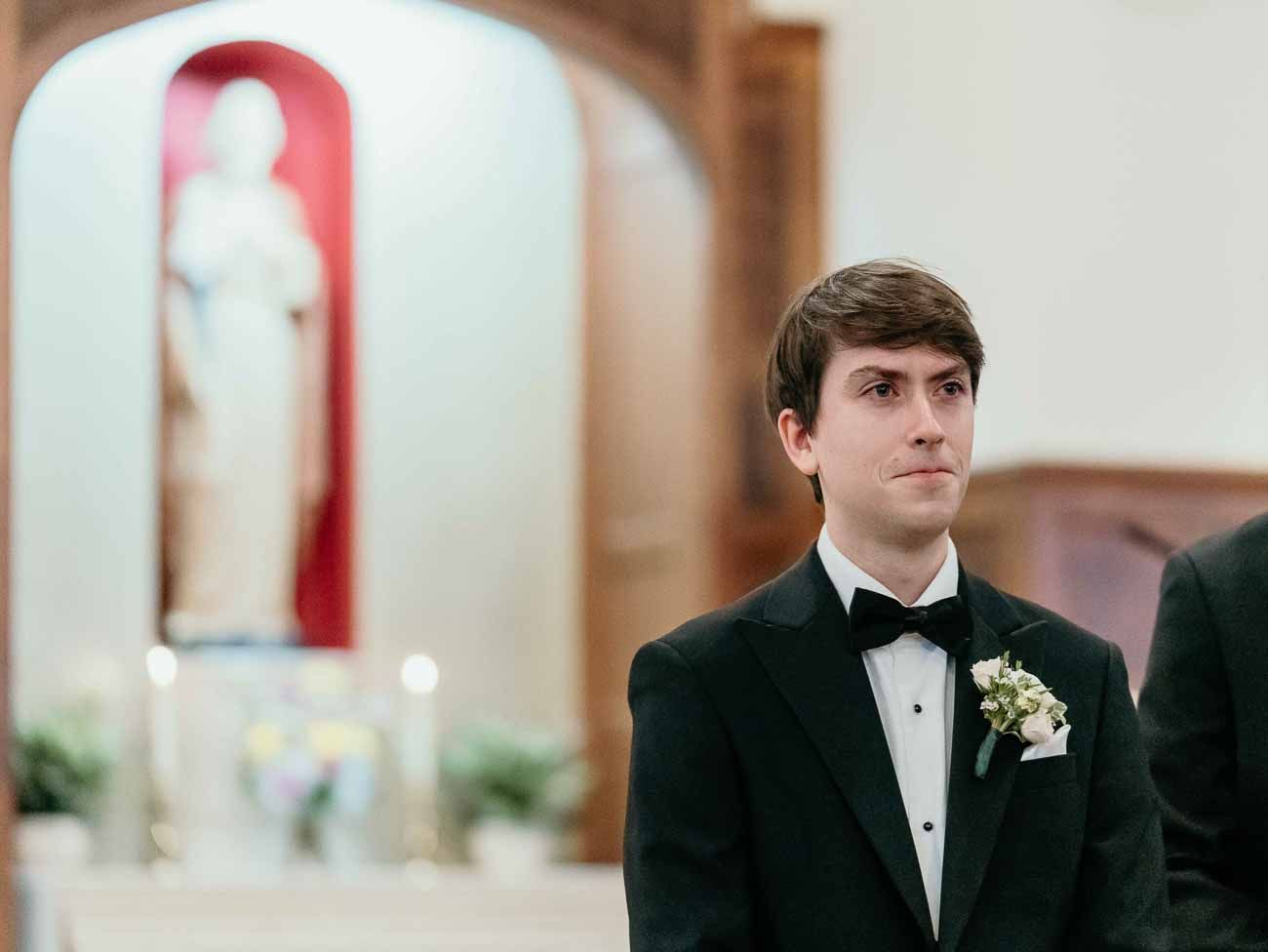 Groom in black tuxedo stands at church altar with a floral boutonniere, awaiting his bride.