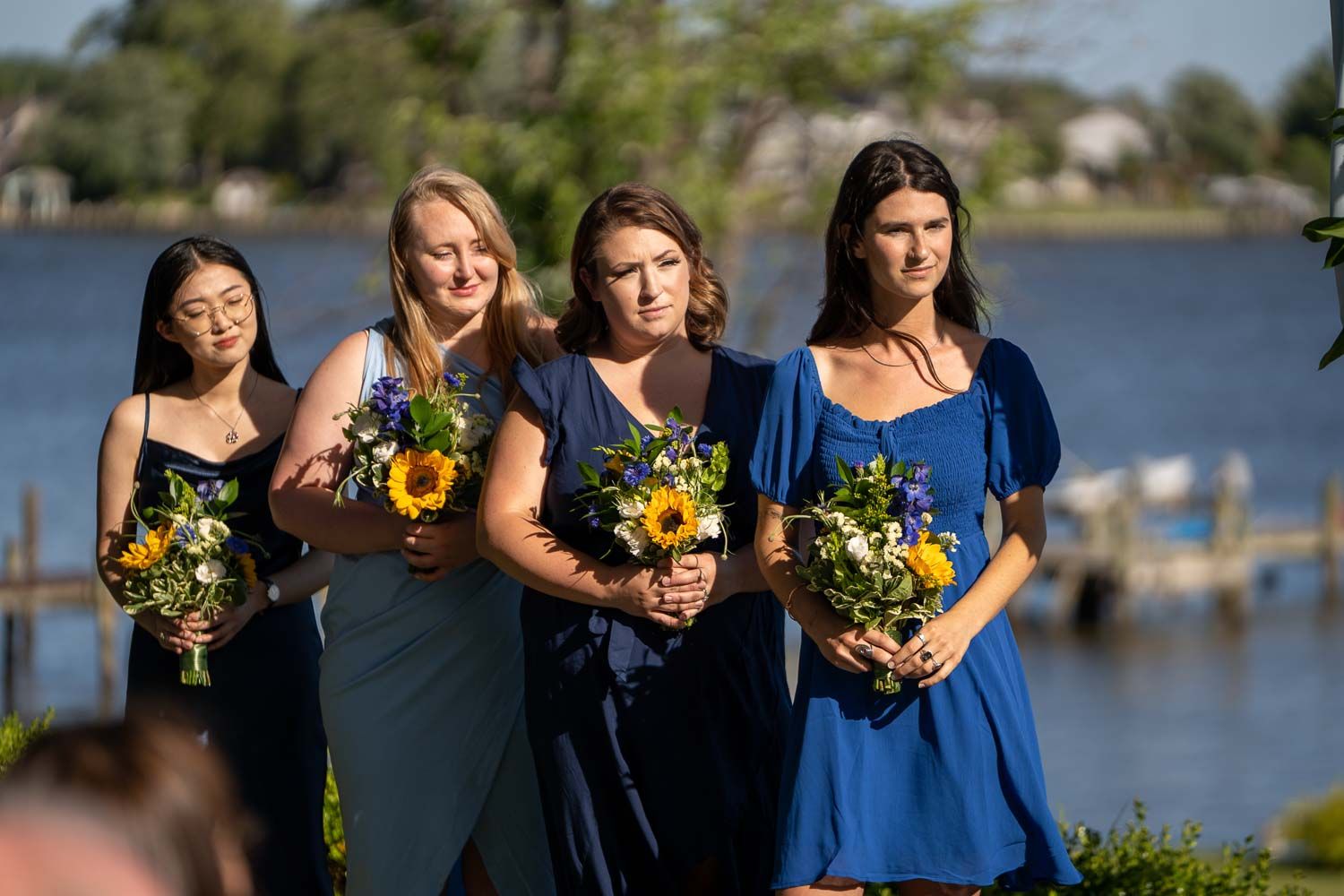 Four bridesmaids in navy blue dresses holding sunflower bouquets stand at waterfront wedding ceremony.