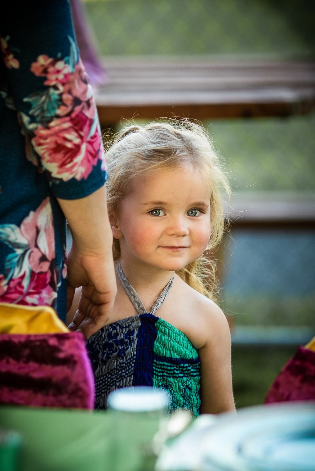 Adorable blonde toddler girl in a teal dress holding an adults hand at an outdoor gathering.