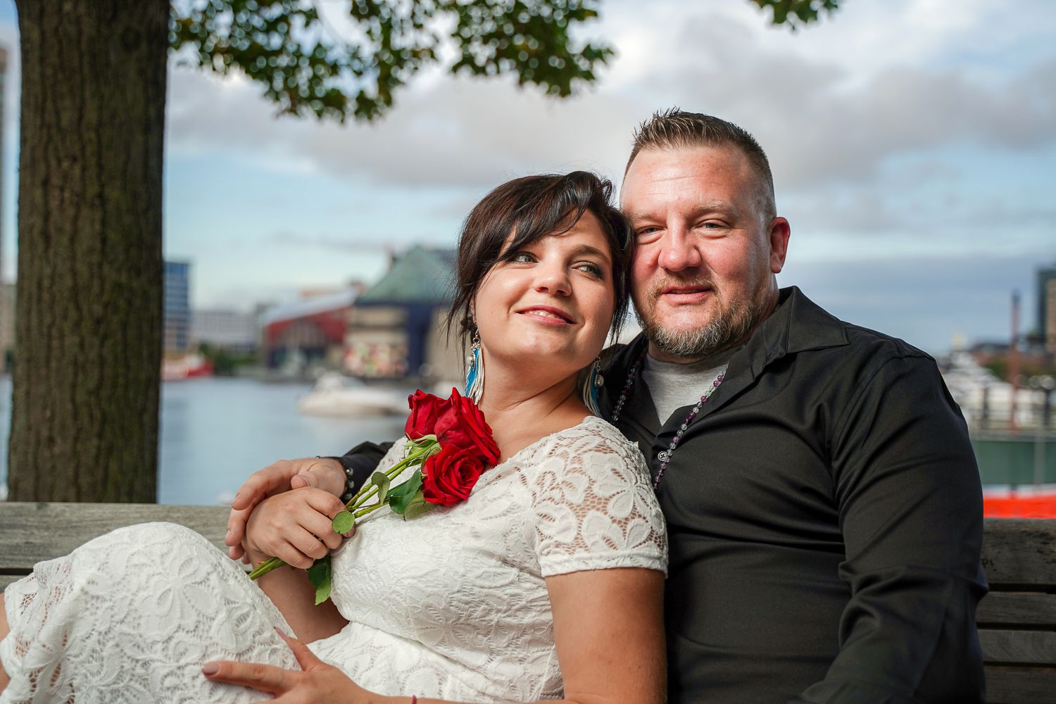 Smiling wedding couple posing on a bench by the harbor, bride in white lace dress with red roses, groom in black shirt.