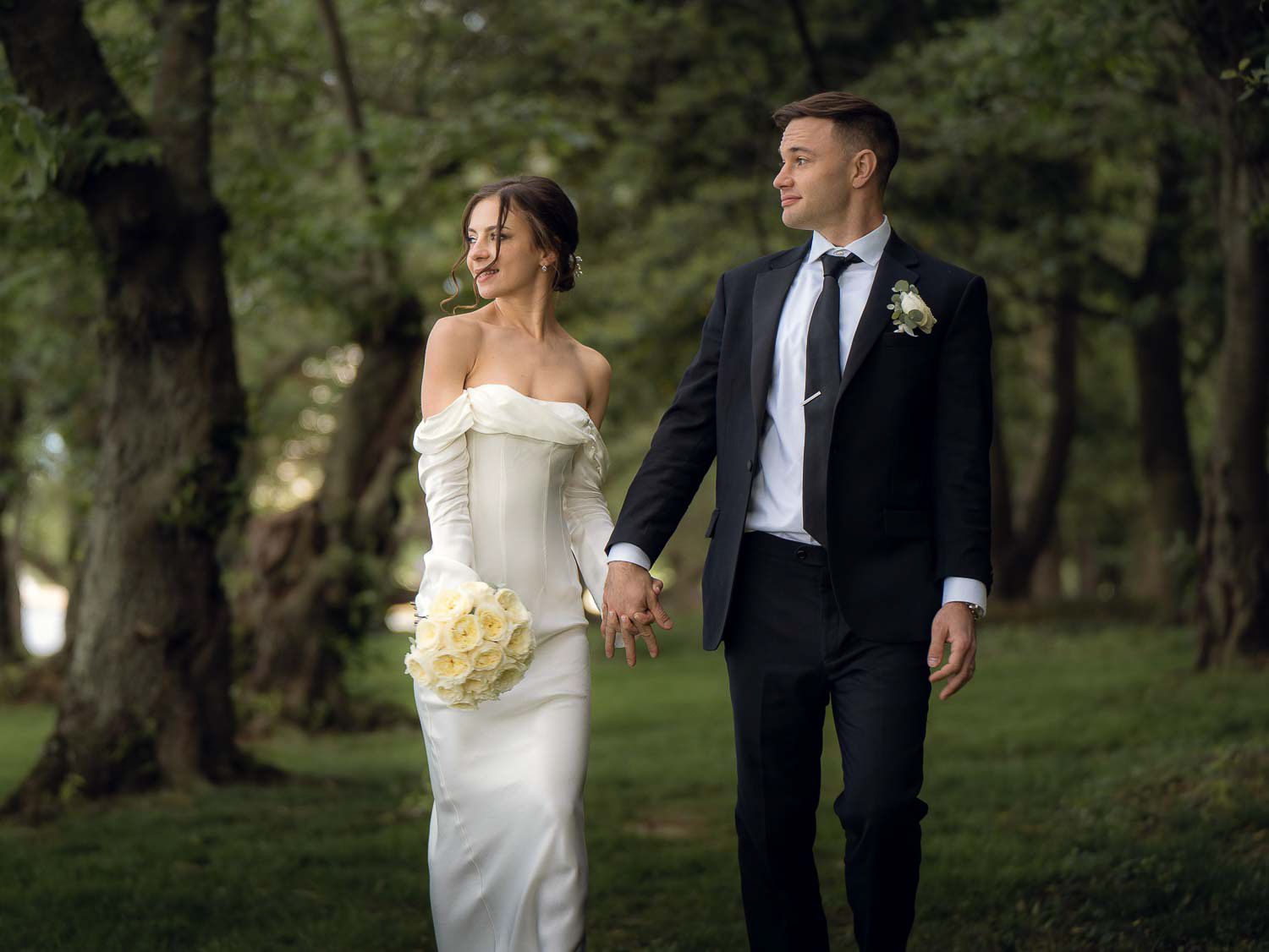 Bride and groom hold hands walking through a sunlit tree-lined park on their wedding day.