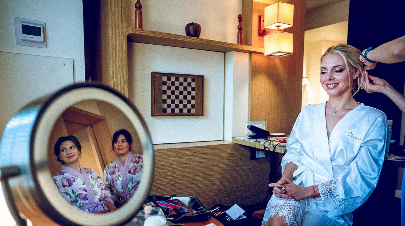 Smiling blonde bride getting hair styled with bridesmaids reflected in mirror.
