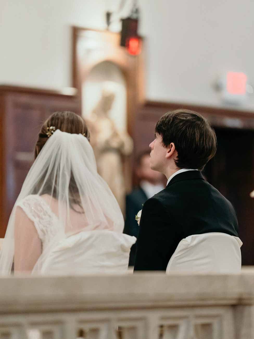 Bride in veil and groom in tuxedo sit together at the altar during Catholic wedding ceremony.