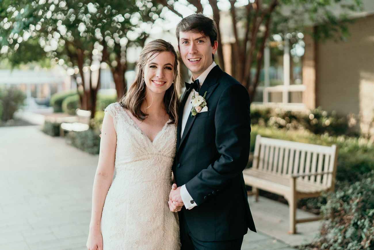 Smiling bride and groom holding hands outdoors on their wedding day, bride in lace gown, groom in black tuxedo.
