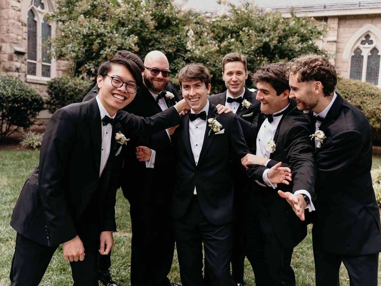 Groomsmen in black tuxedos laugh and celebrate with the groom outside a stone church.