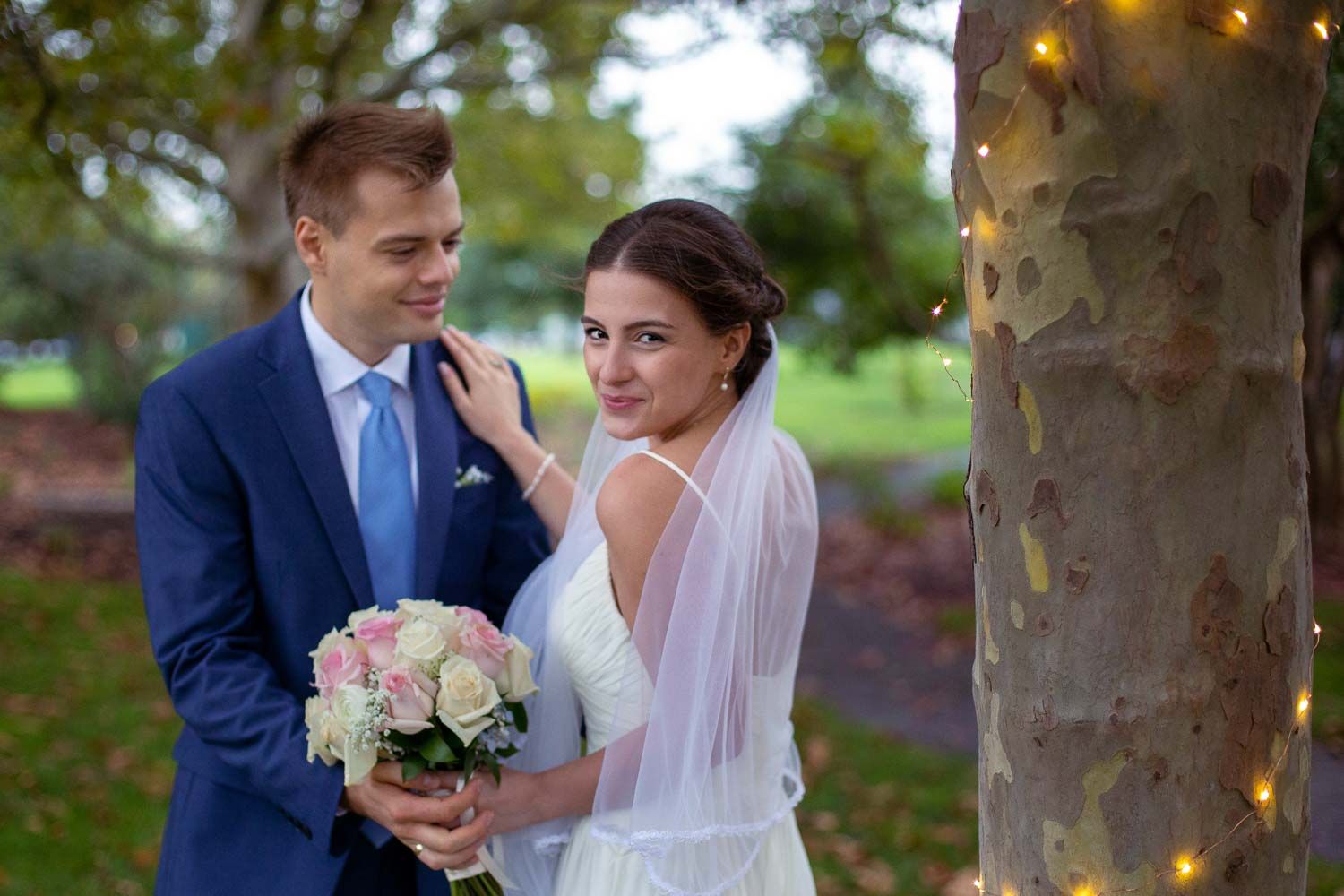 Bride and groom posing outdoors near a tree with fairy lights, bride holding a bouquet of pink roses.
