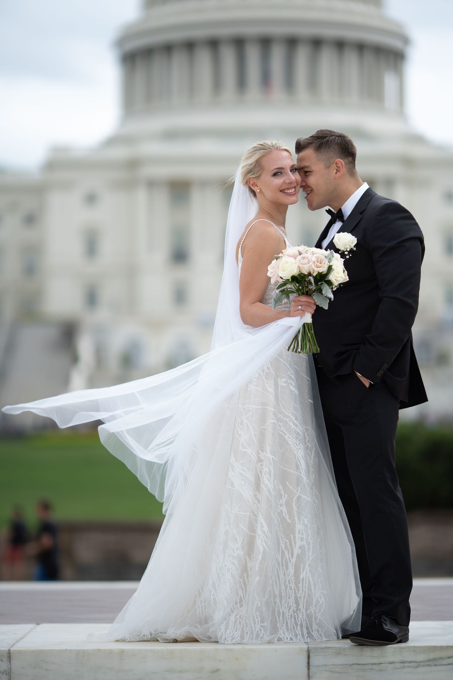 Bride and groom pose romantically in front of the US Capitol building, bride holding white roses bouquet.
