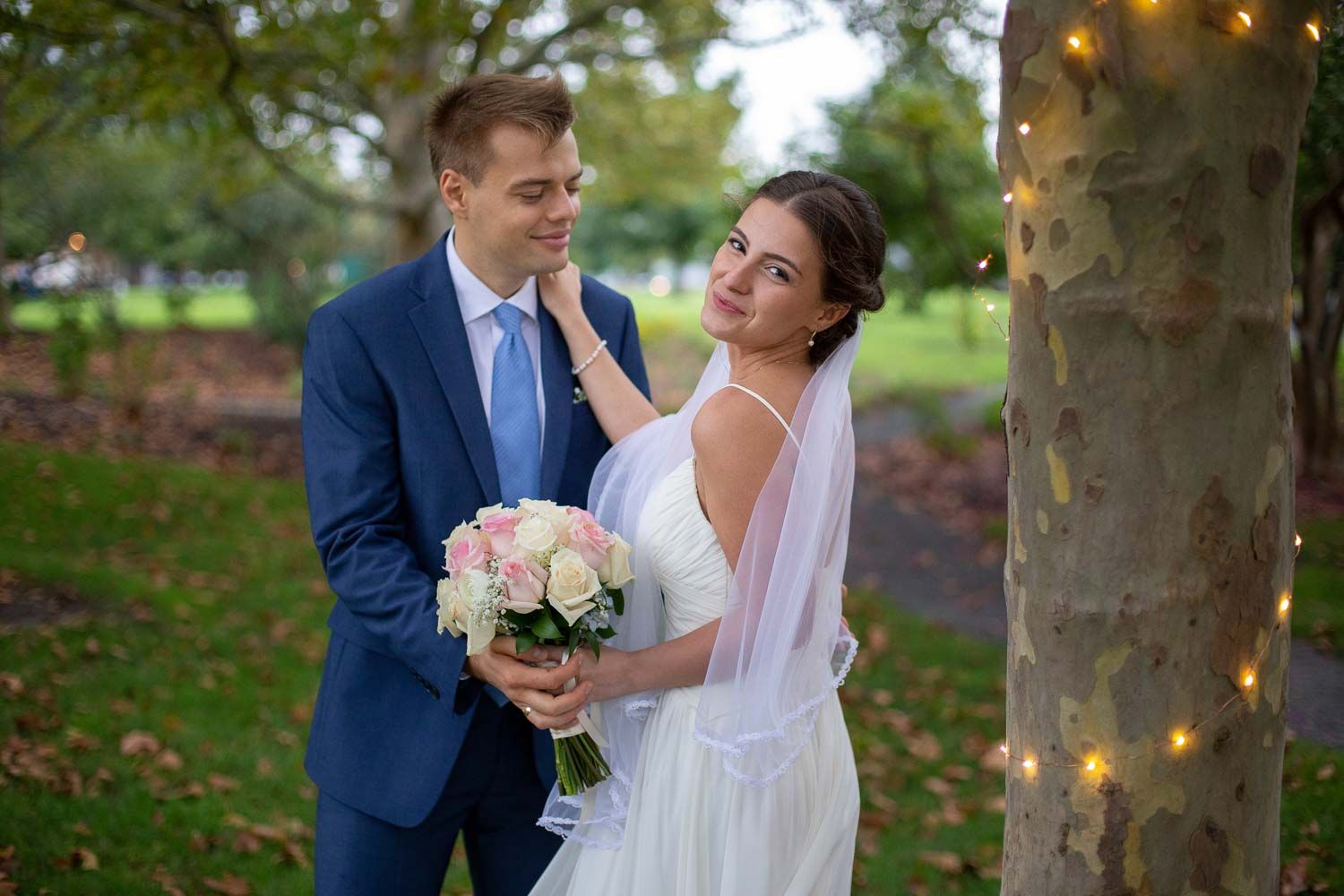 Bride in white dress and veil smiles at groom in navy suit holding pink rose bouquet by a tree.