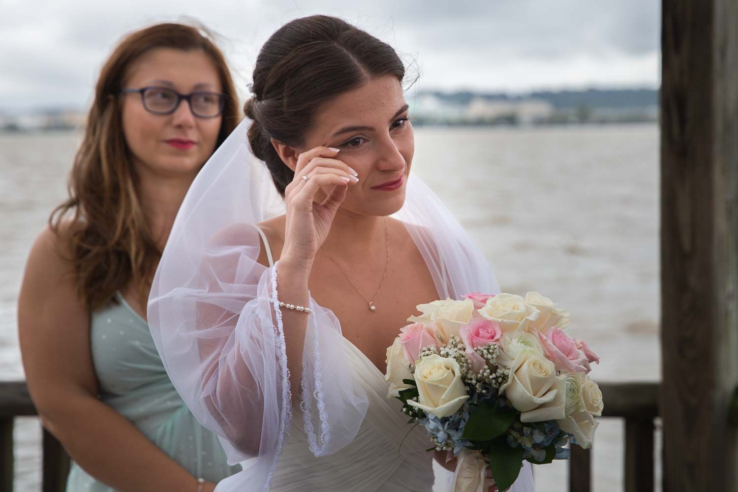 Emotional bride wiping tears while holding a pink and white bouquet with bridesmaid behind her.
