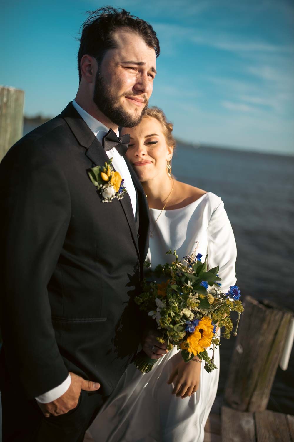 Bride and groom embracing on waterfront dock, bride holding colorful yellow and blue wildflower bouquet in white gown.