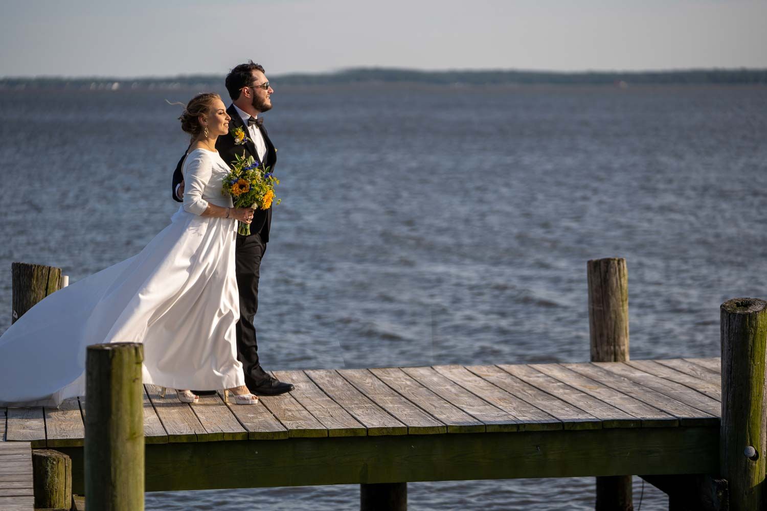 Bride and groom standing on a wooden dock overlooking calm blue water on their wedding day.