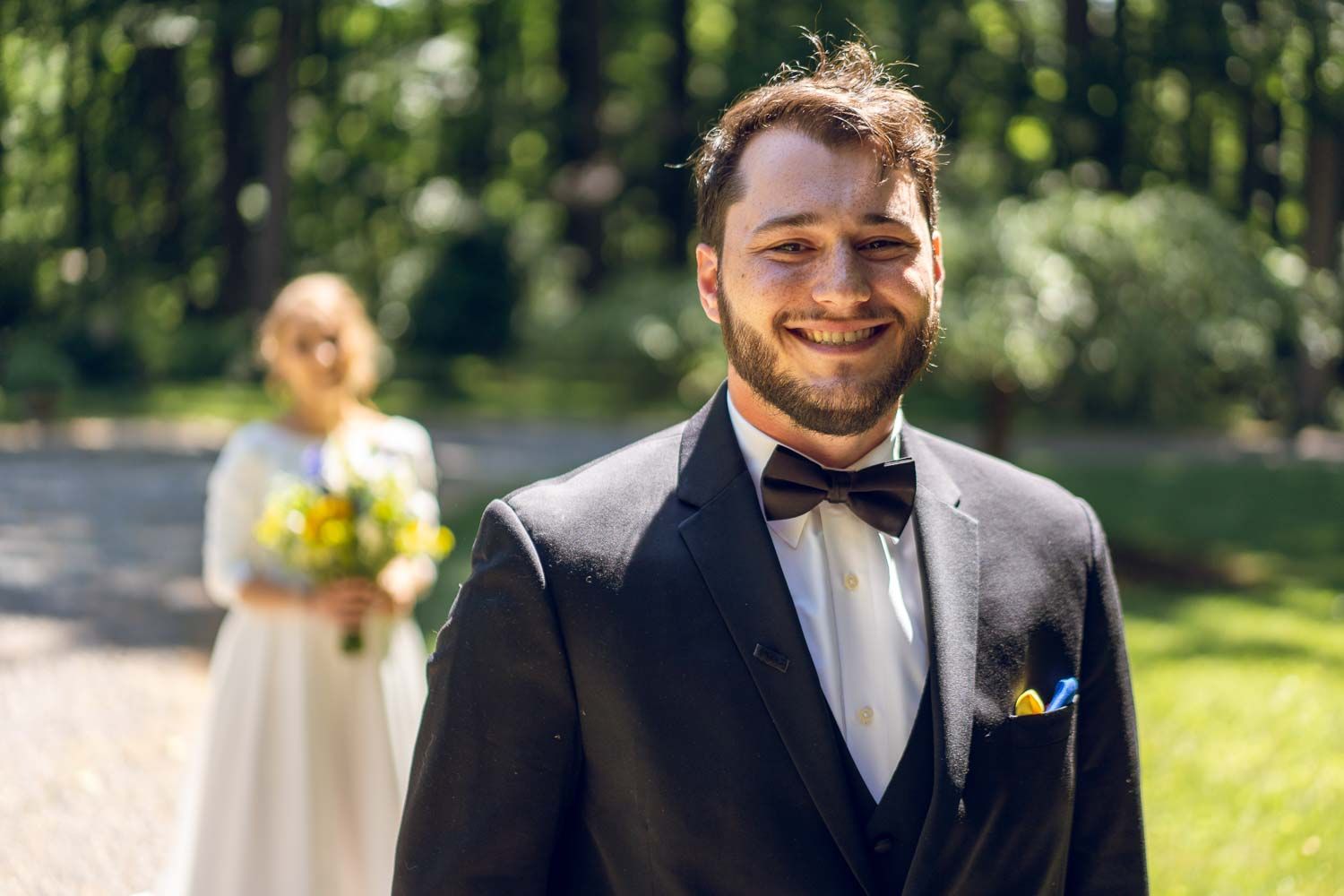 Smiling groom in black tuxedo with bride holding yellow bouquet in background outdoors.