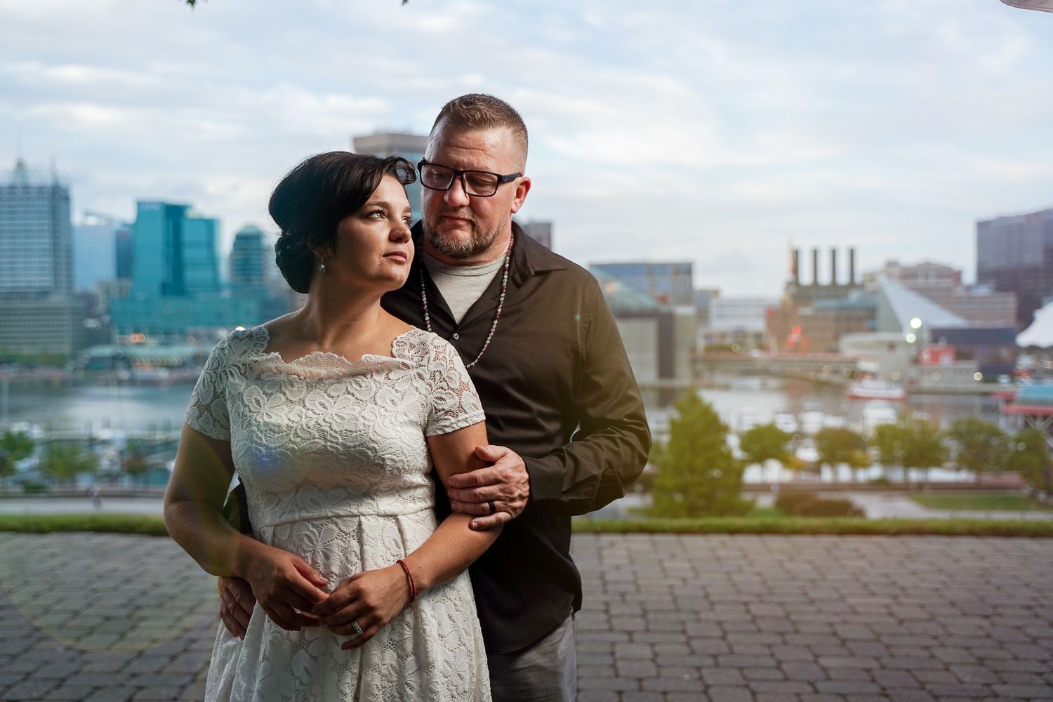 Stylish couple embracing with a scenic harbor and city skyline in the background on a cloudy day.