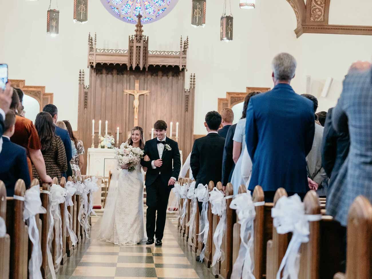 Newlyweds walking down church aisle after wedding ceremony with white ribbon-decorated pews.