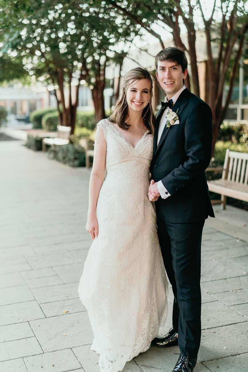 Bride in elegant lace wedding gown and groom in black tuxedo holding hands and posing on a brick pathway.