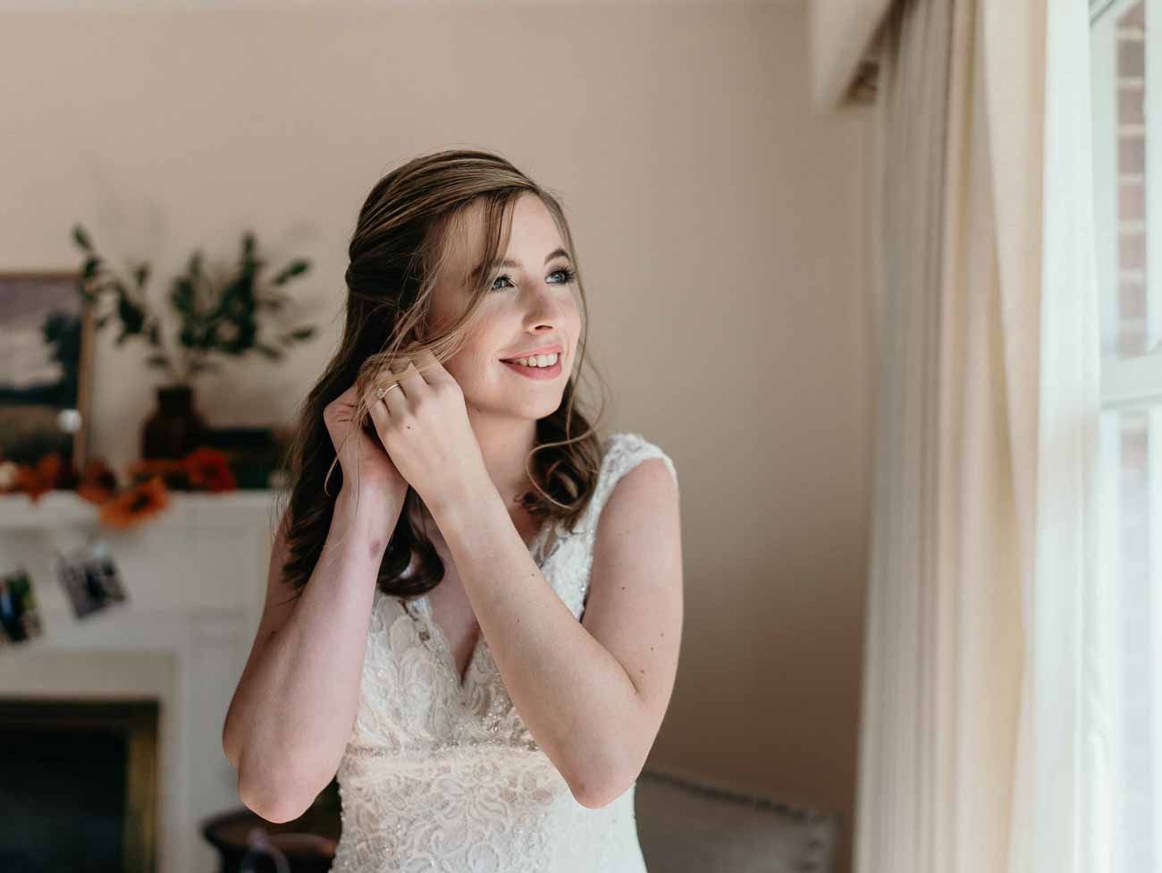 Smiling bride in lace wedding dress adjusts earring near bright window on wedding day.