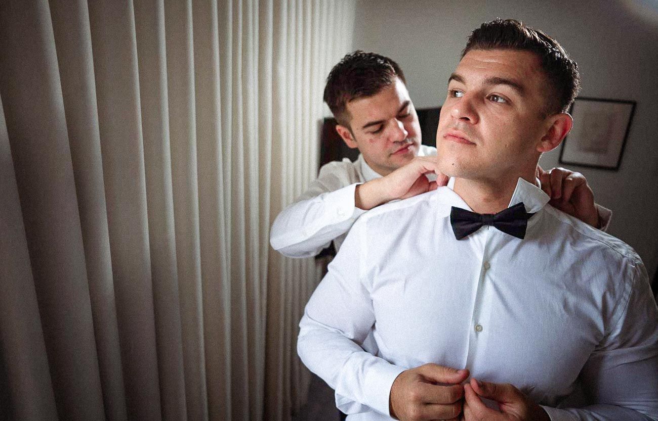 Groomsman helping groom fix bow tie while getting ready for wedding.