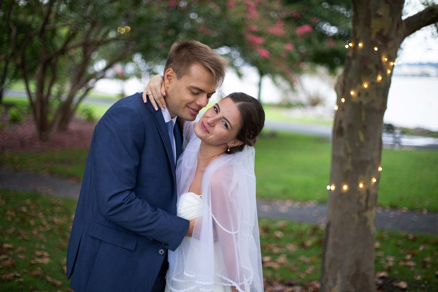 Groom in navy suit embracing smiling bride in white dress and veil near a tree with fairy lights.