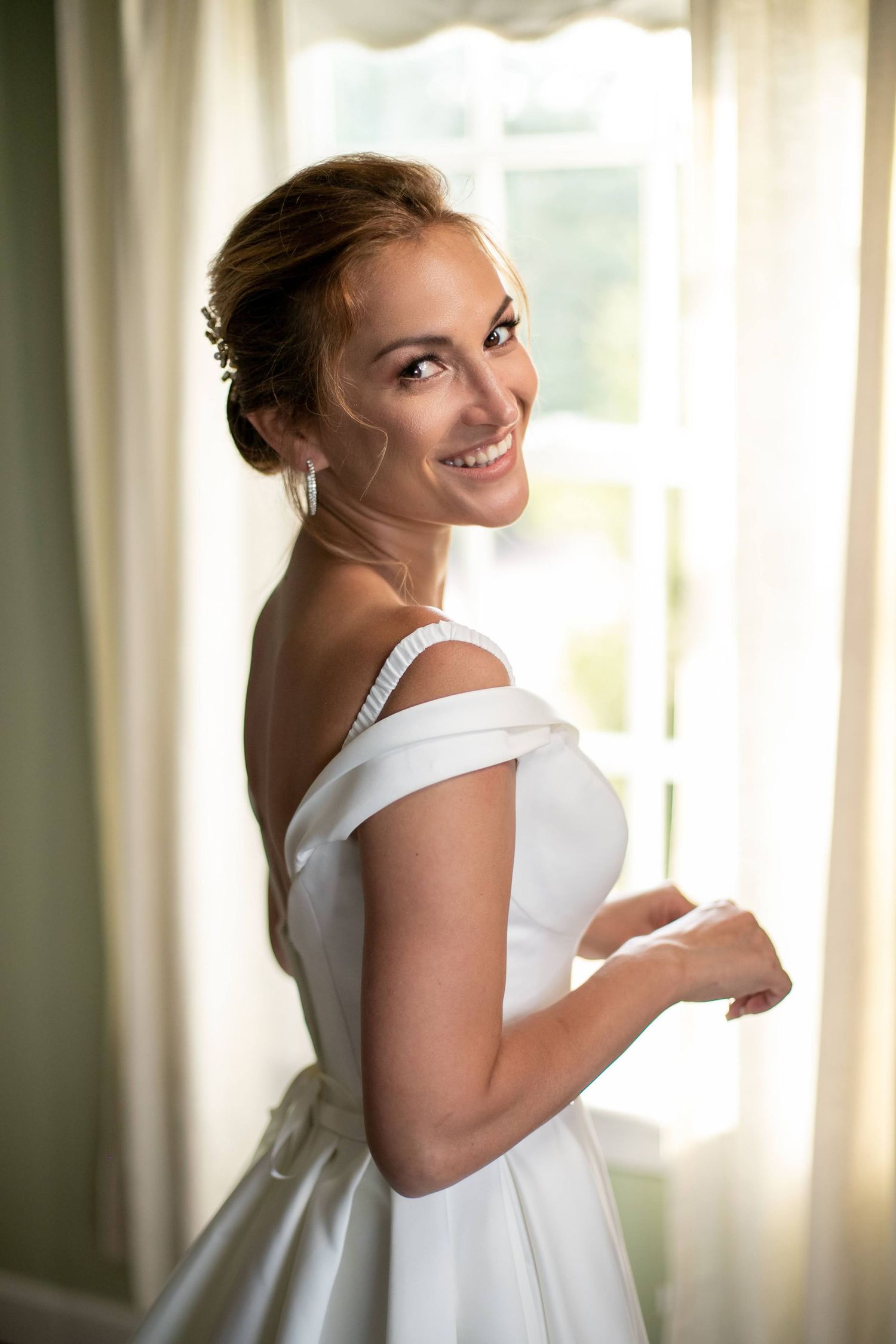 A smiling bride in an off-shoulder white satin gown stands elegantly by a sunlit window.
