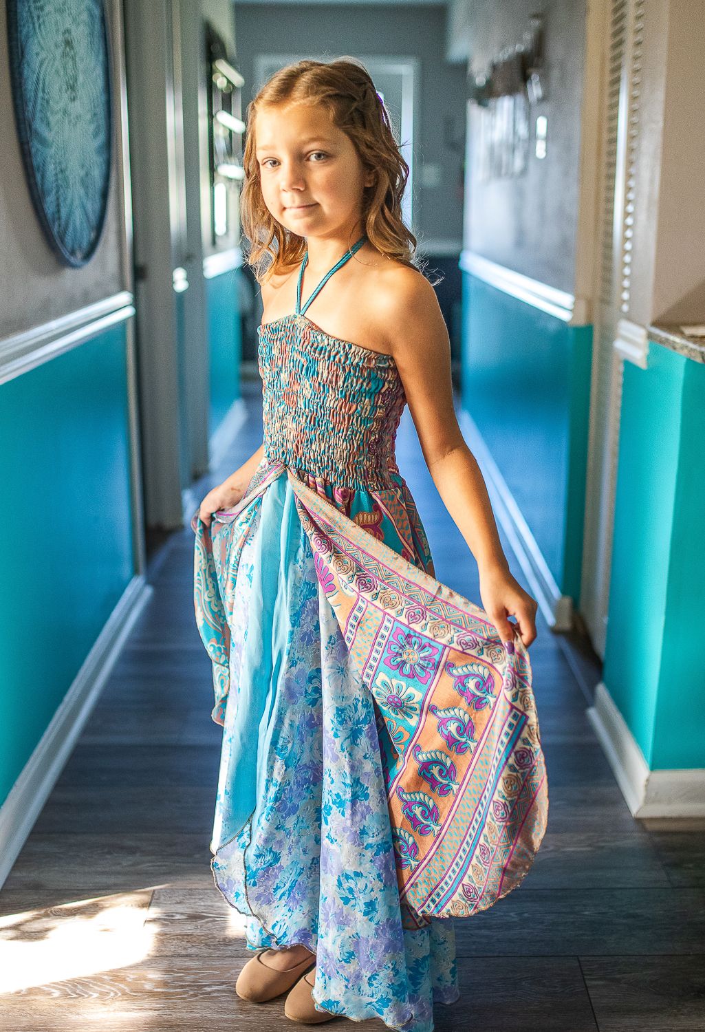 Young girl twirling in a vibrant blue floral maxi dress in a sunlit teal hallway.