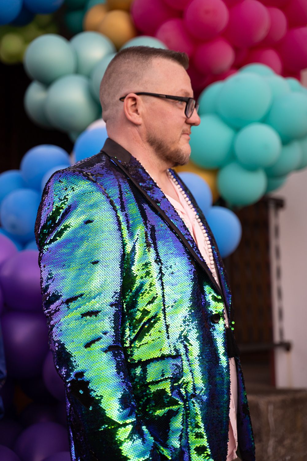 Man in stunning iridescent sequin blazer poses in front of colorful balloons at festive outdoor celebration.