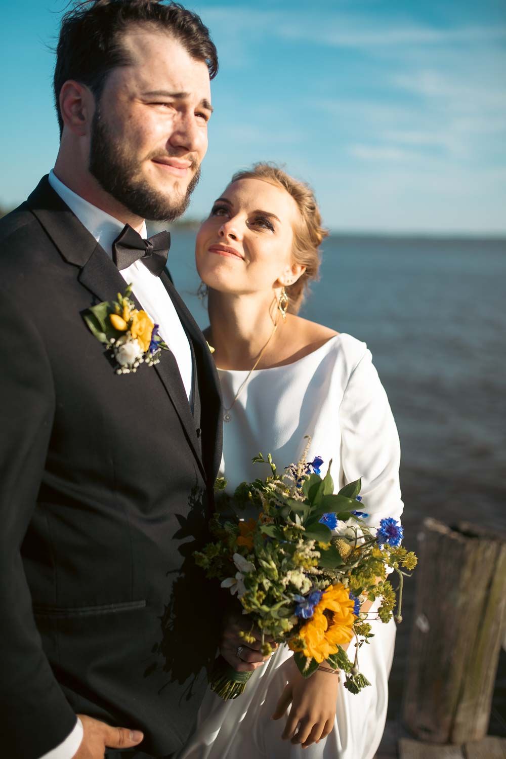 Bride in white dress leaning on groom in tuxedo, holding vibrant sunflower and blue wildflower bouquet by the sea.