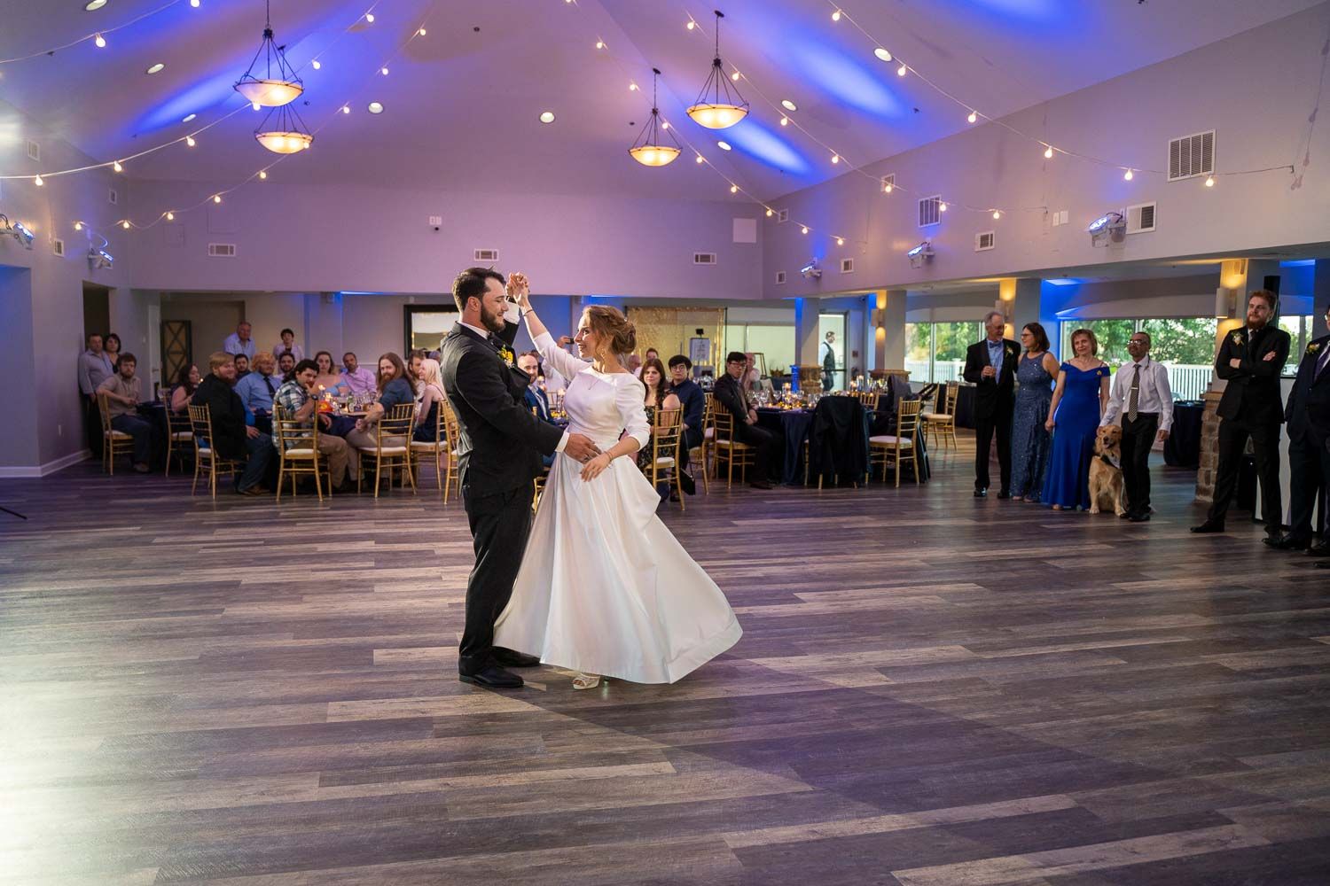 Newlyweds sharing first dance in elegant reception venue with blue uplighting and vaulted ceiling.