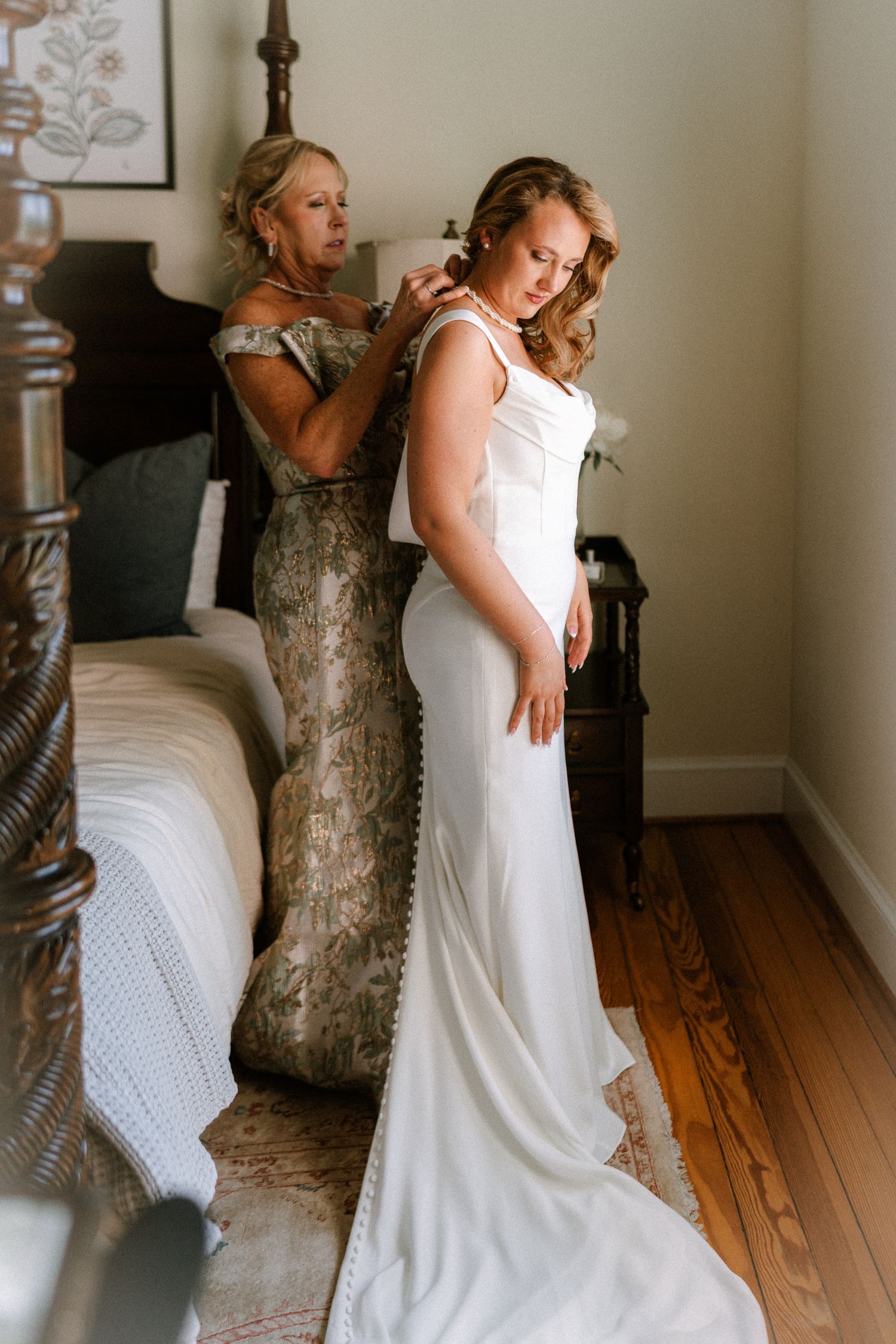 Mother helps bride in elegant white gown with necklace in a warmly lit bedroom before wedding ceremony.