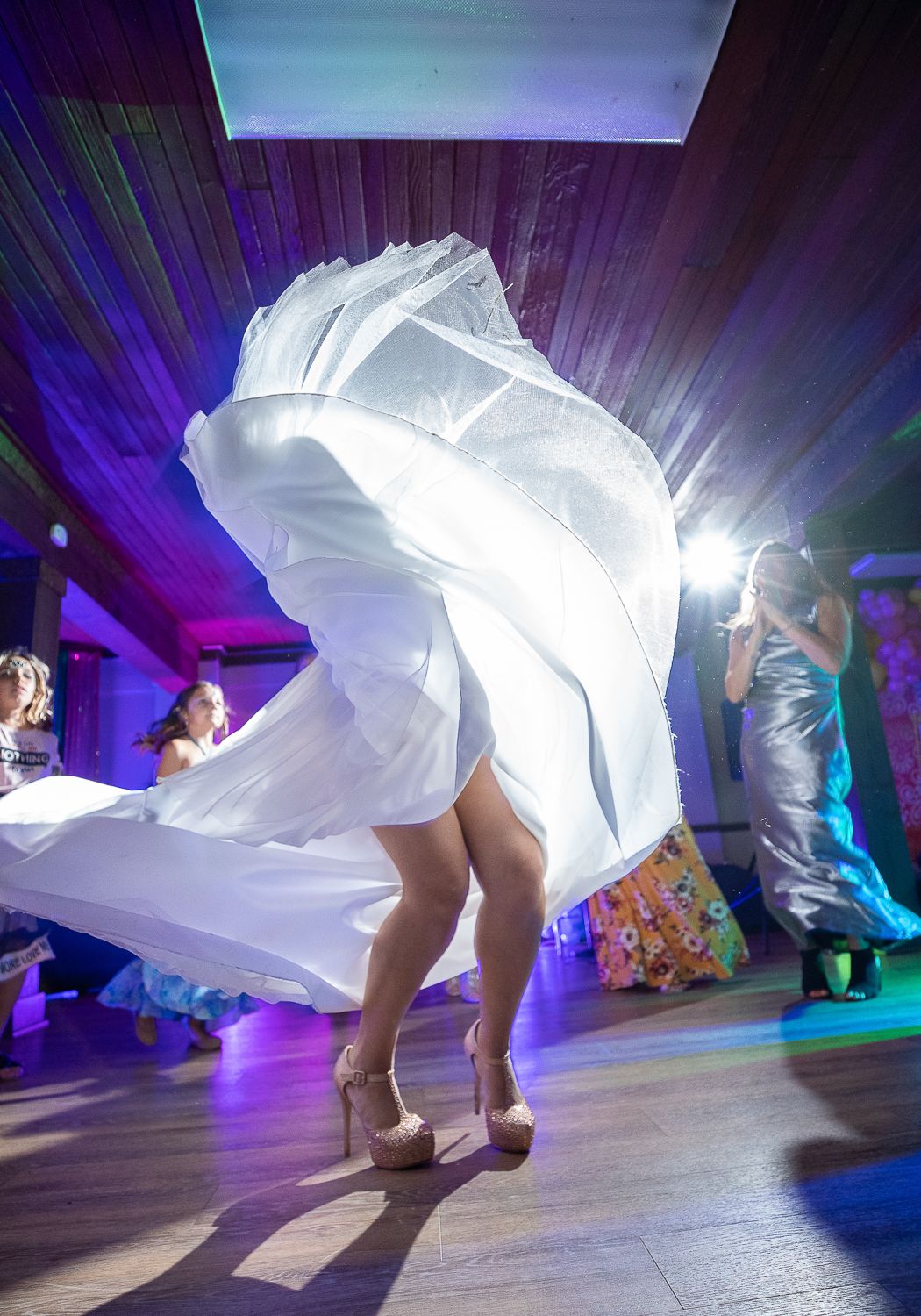 Bride twirling in white wedding dress on dance floor with colorful lights at wedding reception.
