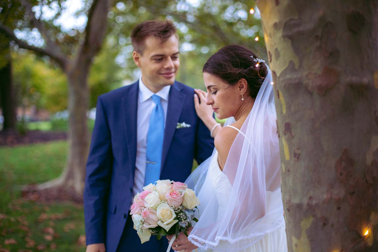Groom in navy suit gazes at emotional bride holding pink rose bouquet near a tree in a park.