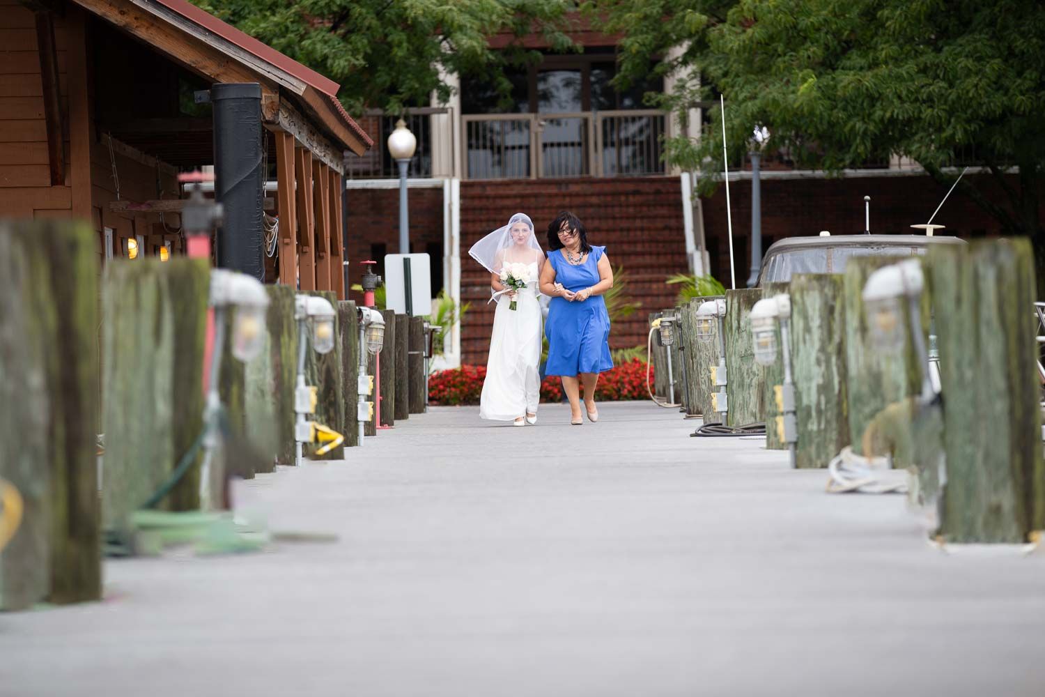 Bride in white wedding dress walking along a marina dock accompanied by a woman in blue dress.