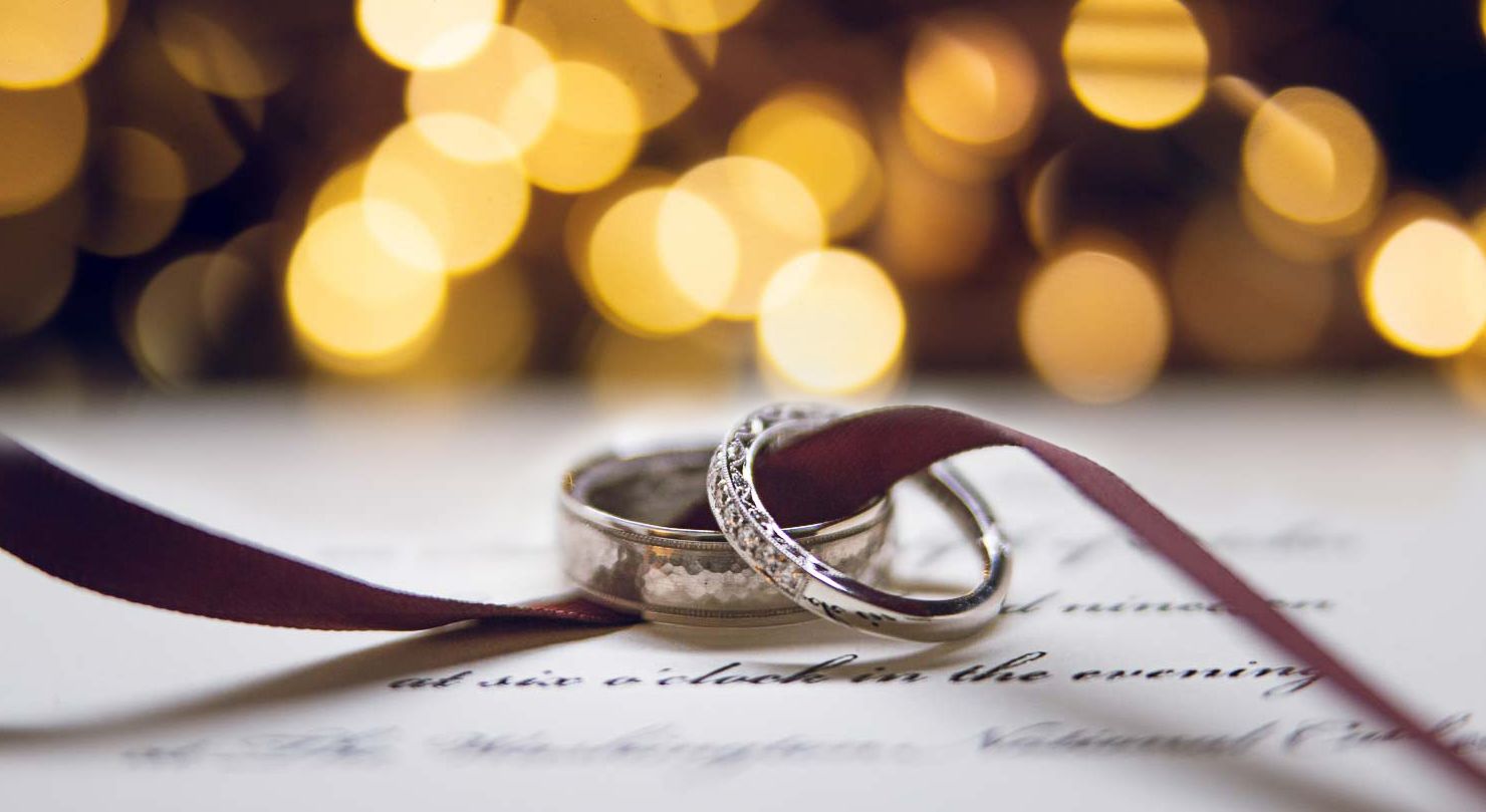 Close-up of two elegant silver wedding rings tied with a burgundy ribbon on a written page with bokeh lights.