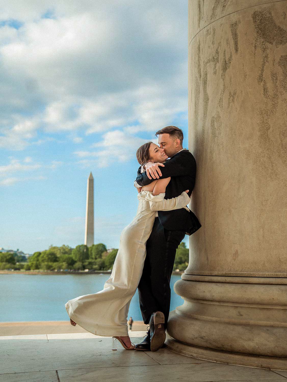 Couple embracing against marble column with Washington Monument in background.