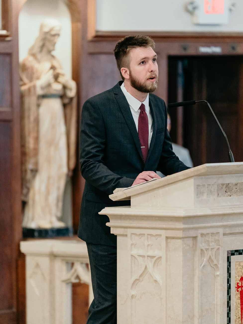 Man in dark suit delivers reading at ornate marble church podium during wedding ceremony.