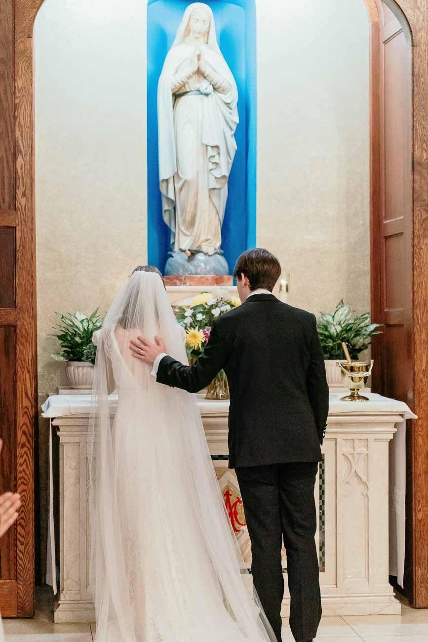 Bride and groom praying before Virgin Mary statue at church side altar during wedding ceremony.