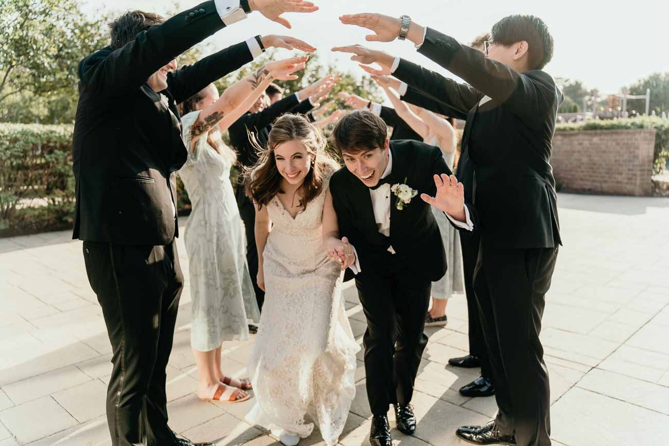 Bride and groom duck under an arch of raised hands formed by their wedding party outdoors.