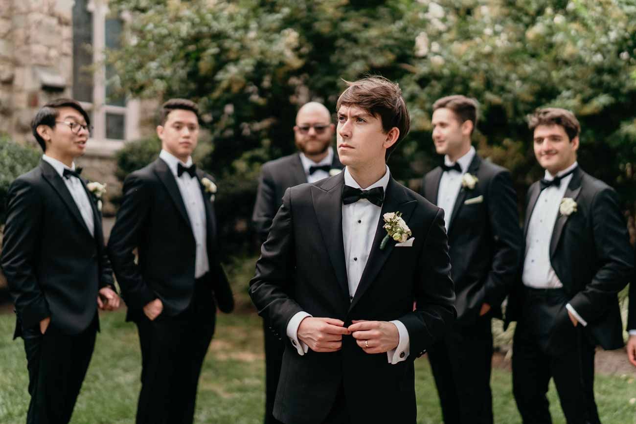 Groom in black tuxedo with floral boutonniere stands confidently before his groomsmen outdoors.