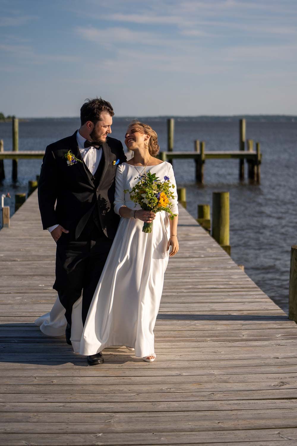 Smiling bride and groom walking along wooden waterfront dock on sunny wedding day.